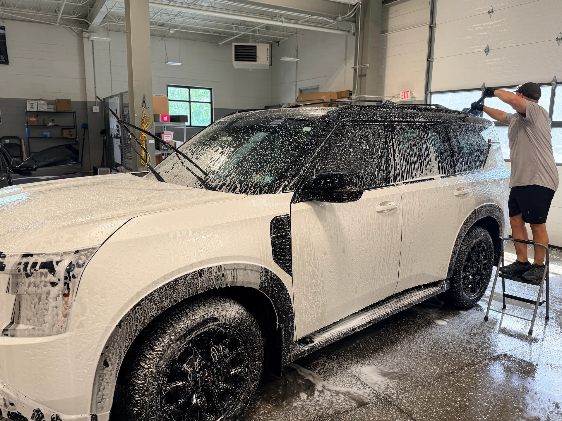 A white SUV covered in foam during a hand wash, with a technician working the roof from a step stool.